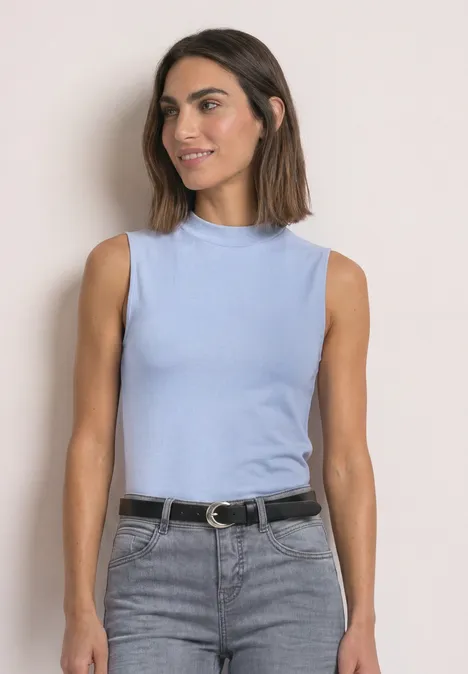 Basic top cashmere blue Frau in ärmellosem hellblauen Top und grauer Jeans mit schwarzem Gürtel vor beigem Hintergrund, seitlich lächelnd.