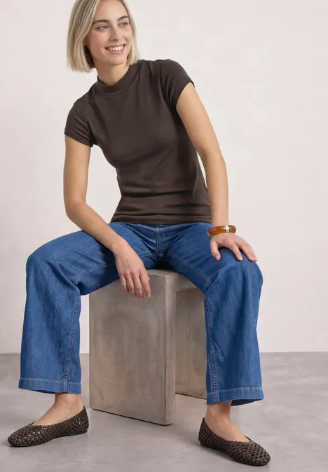Basic Shirt mit Turtle Neck espresso brown Frau mit blondem Bob sitzt auf einem Block, trägt braunes T-Shirt, weite Jeans, braune geflochtene Ballerinas und Holzarmreif.