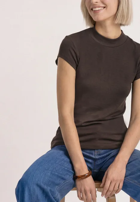 Basic Shirt mit Turtle Neck espresso brown Frau sitzt lächelnd, trägt braunes T-Shirt, blaue Jeans und Holzarmband.