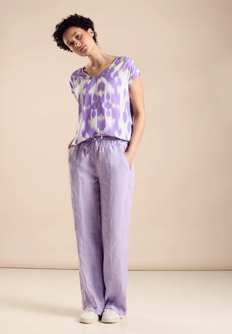 Viskose Bluse smell of lavender Frau in lila Musterbluse und lockerer Hose, Hände in den Taschen, heller Hintergrund.