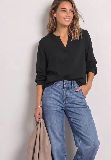 Split Neck Tunika Black Frau mit schwarzer Bluse und weiter blauer Jeans hält eine Jacke und lächelt.