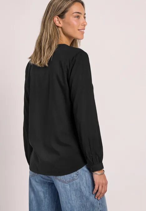 Split Neck Tunika Black Frau von hinten in schwarzer Bluse und blauen Jeans, mit leicht gedrehtem Kopf Richtung Betrachter.