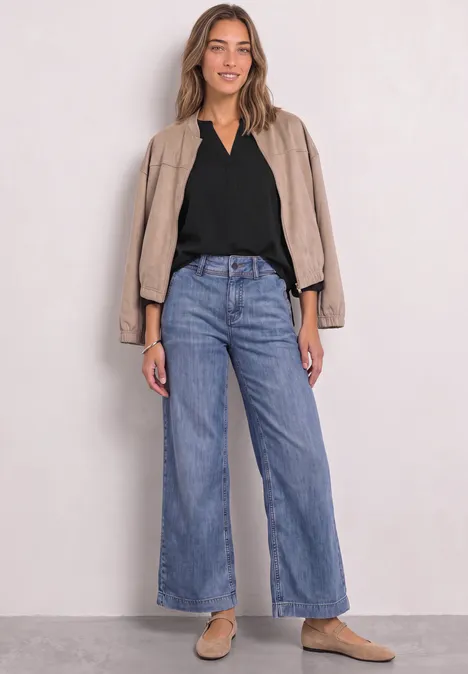 Split Neck Tunika Black Frau in lockerer Jeans mit weitem Bein, schwarzem Top und beiger Jacke, lässig mit hellen Slippern.