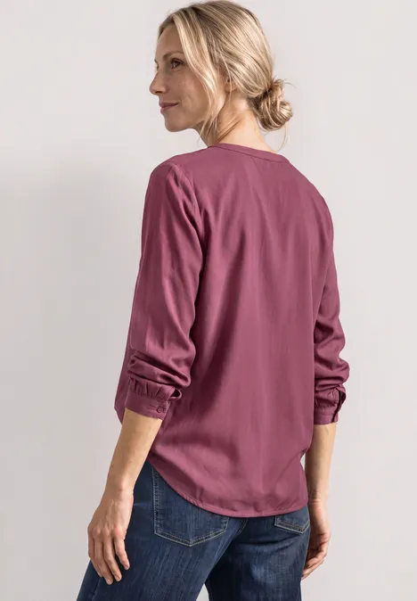 Split Neck Tunika dark clouded blush Frau im Profil trägt eine lachsrosa Bluse mit hochgekrempelten Ärmeln und Jeans auf hellem Hintergrund.