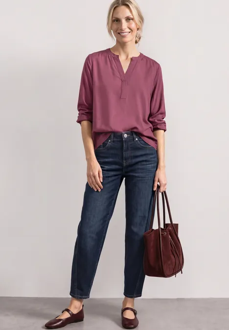 Split Neck Tunika dark clouded blush Eine Frau vor neutralem Hintergrund trägt dunkle Jeans, eine plumfarbene Bluse und eine große Handtasche.