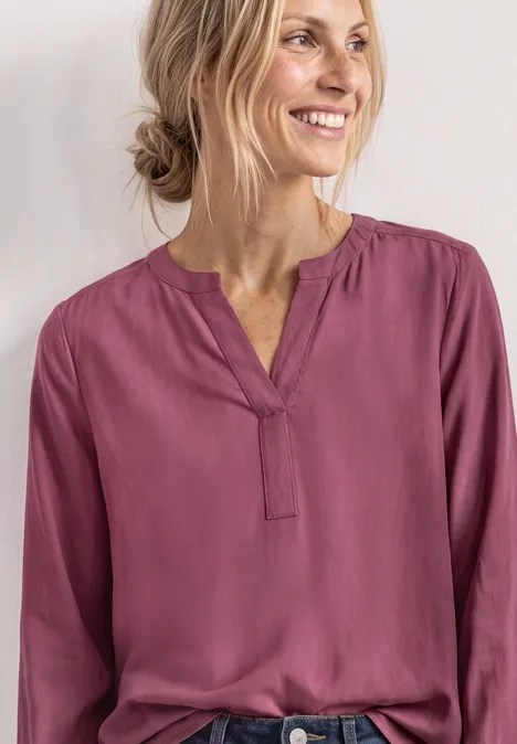 Split Neck Tunika dark clouded blush Eine lächelnde Frau in einer violetten, locker sitzenden Bluse vor hellem Hintergrund.