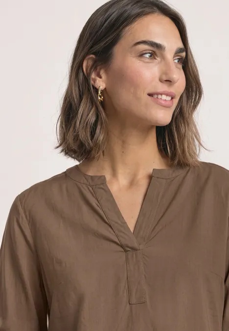 Tuniek met V-hals sanded amber Porträt einer lächelnden Frau mit brauner Bluse und goldenen Ohrringen, seitlich blickend.