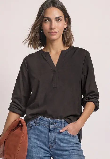 Split Neck Tunika black coffee Frau in schwarzer Bluse mit V-Ausschnitt, hochgekrempelten Ärmeln und Jeans, die eine braune Handtasche hält.