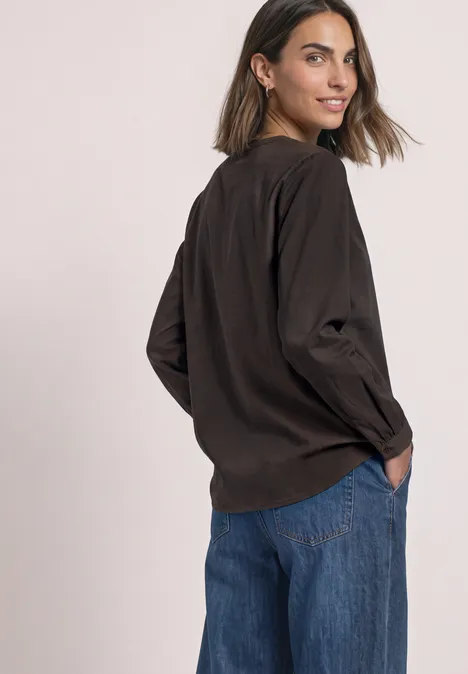 Split Neck Tunika black coffee Frau in dunkler Bluse und blauen Jeans dreht sich leicht zur Kamera, Hände in den Taschen.