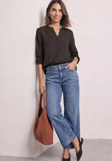 Split Neck Tunika black coffee Frau in schwarzem Langarmshirt, blauen Jeans und brauner Tasche lehnt an weißer Wand.