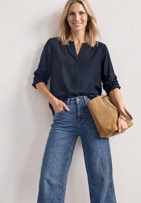 Frau in dunkelblauer Bluse und weitem Jeans, mit brauner Tasche, steht lächelnd vor heller Wand.