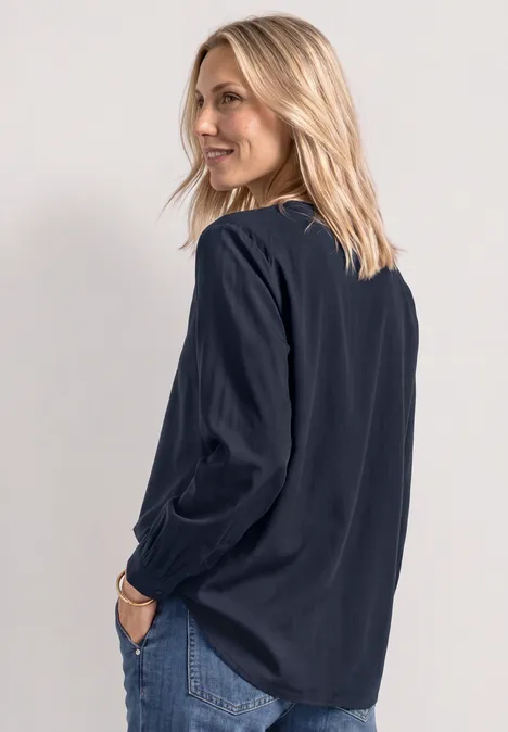 Eine Frau mit blonden Haaren trägt eine dunkelblaue Bluse und Jeans, schaut lächelnd über die Schulter.