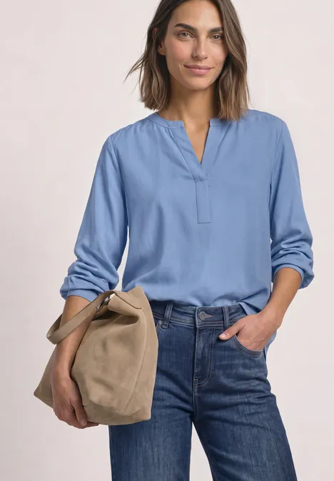 Vrouw in lichtblauw shirt en jeans met een beige tas voor een effen achtergrond.