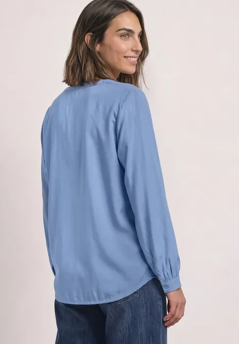 Vrouw met lichtblauw blouse met lange mouwen en spijkerbroek kijkt glimlachend over haar schouder.