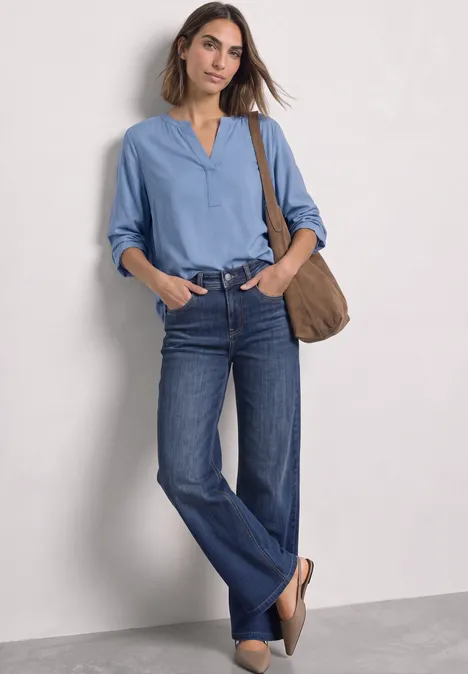 Vrouw in lichtblauw overhemd, blauwe jeans en tas, staat nonchalant voor een neutrale achtergrond.