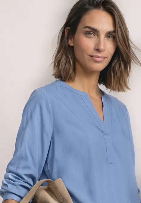 Een lachende vrouw met halflang haar draagt een lichtblauwe tuniek en houdt een beige handtas vast.