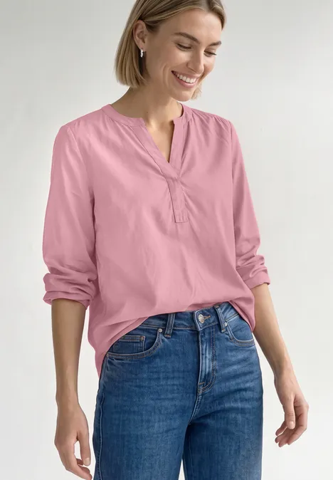 Tuniek met V-hals vintage blush Eine Frau trägt eine rosa Bluse und blaue Jeans, lächelt und posiert entspannt.