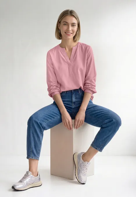 Tuniek met V-hals vintage blush Eine Frau sitzt lächelnd auf einem Würfel, trägt pinke Bluse, blaue Jeans und weiße Sneaker vor weißem Hintergrund.