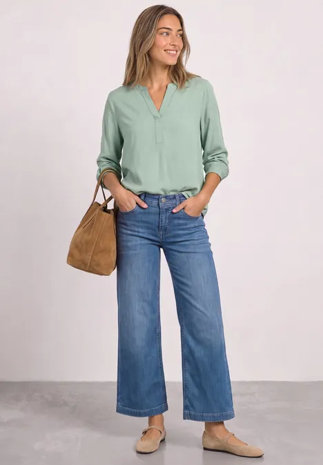 Eine lächelnde Frau in grüner Bluse, weiten Jeans, hellen Schuhen und brauner Tasche vor neutralem Hintergrund.