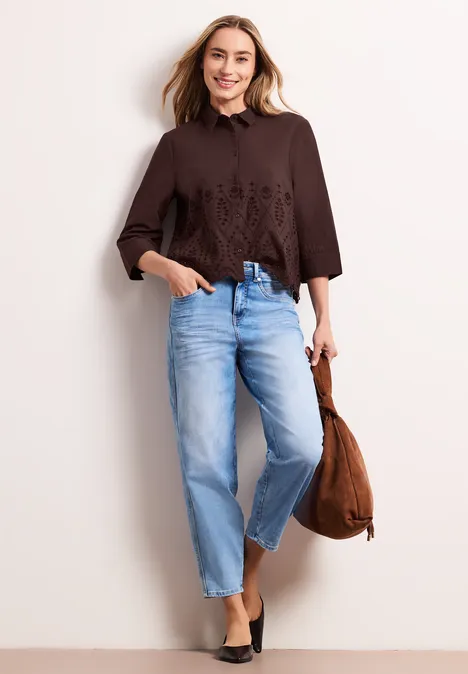 Frau in brauner, bestickter Bluse, hellblauen Jeans, schwarzen Schuhen und brauner Tasche.