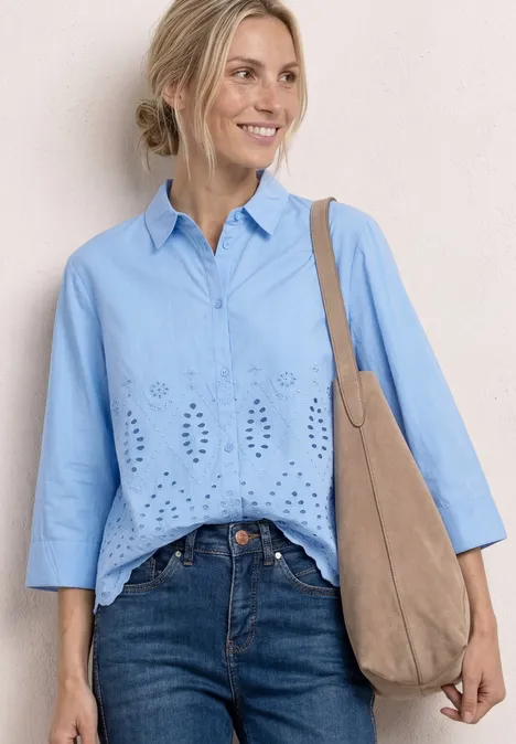 Vrouw in lichtblauwe blouse met open borduursels, spijkerbroek en beige suède tas voor neutrale achtergrond.