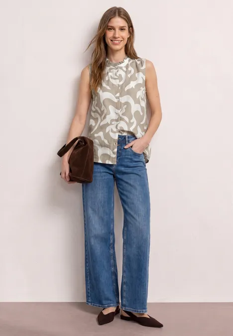 Vrouw in mouwloos dessin/top, rechte jeans, zwarte ballerina’s en bruine handtas.