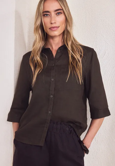 Person in olivgrünem Hemd mit 3/4-Ärmeln und dunkler Hose vor weißer Wand.