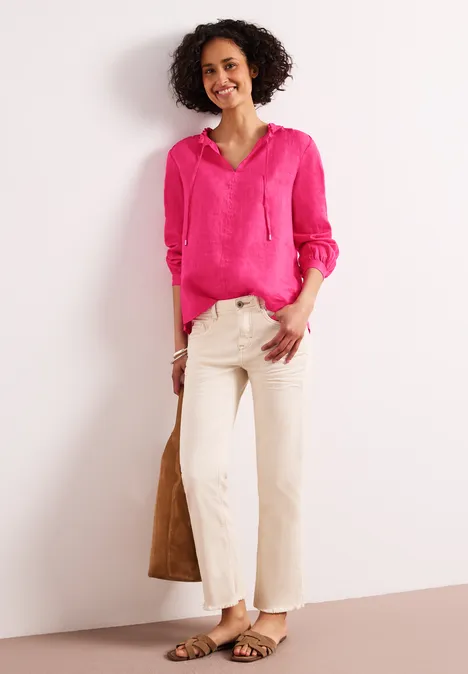 Frau mit pinker Bluse, beiger Hose, Sandalen und brauner Tasche vor weißem Hintergrund.