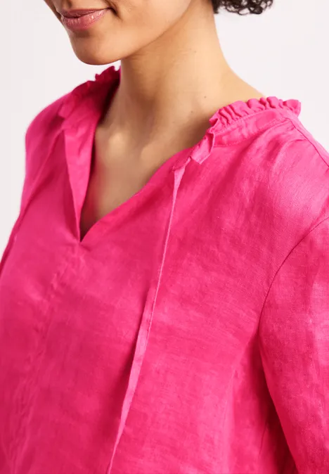 Eine Person trägt eine leuchtend pinke Leinenbluse mit V-Ausschnitt und Rüschenkragen.