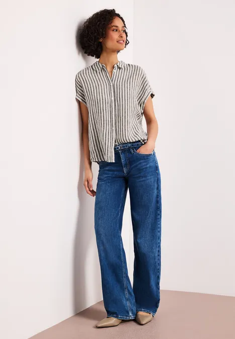 Eine Person lehnt an einer Wand, trägt gestreiftes Hemd und weite blaue Jeans.