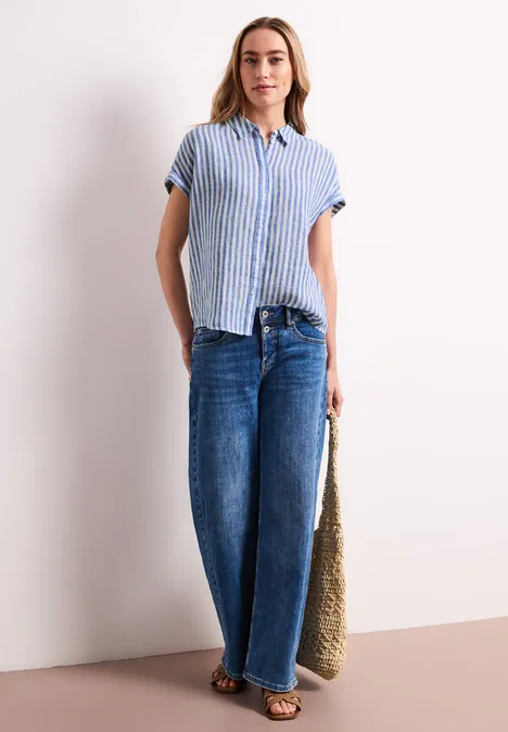 Vrouw in blauw-wit gestreept overhemd met korte mouwen, wijde jeans, sandalen en grote groene tas.