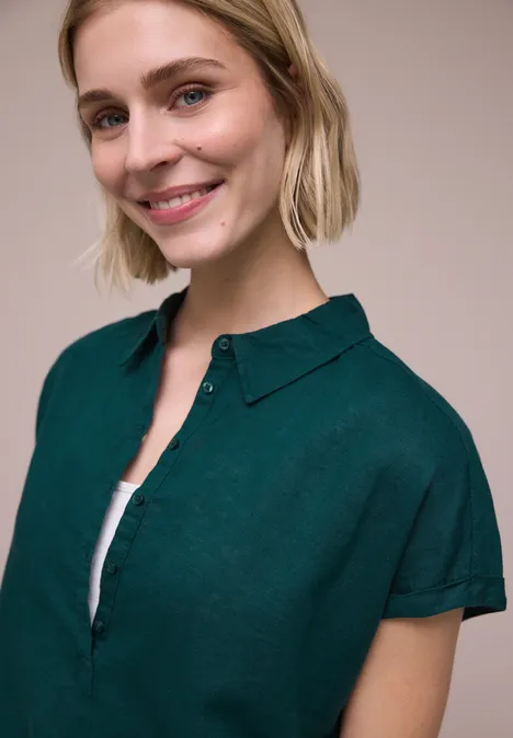 Iemand met blond haar draagt een donker groen kort mouw overhemd op een witte top, gezicht onscherp.