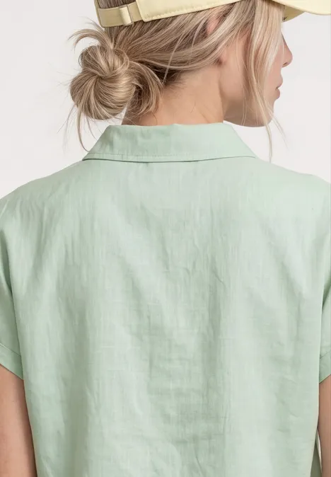 Achteraanzicht van een vrouw met losse knot en baseballpet, die een mintgroen linnen overhemd met korte mouwen draagt.