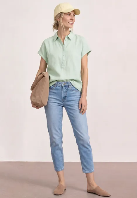 Vrouw in casual zomerse outfit: lichtgroene blouse, blauwe jeans, beige loafers en een tas.