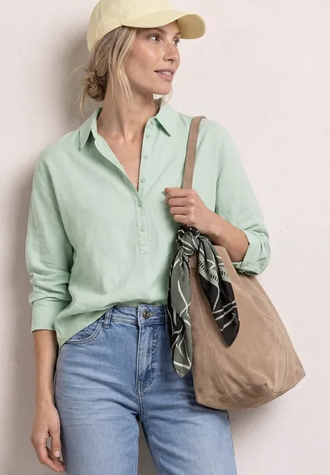 Vrouw in licht linnen overhemd, spijkerbroek, pet en stoffen tas met sjaal.