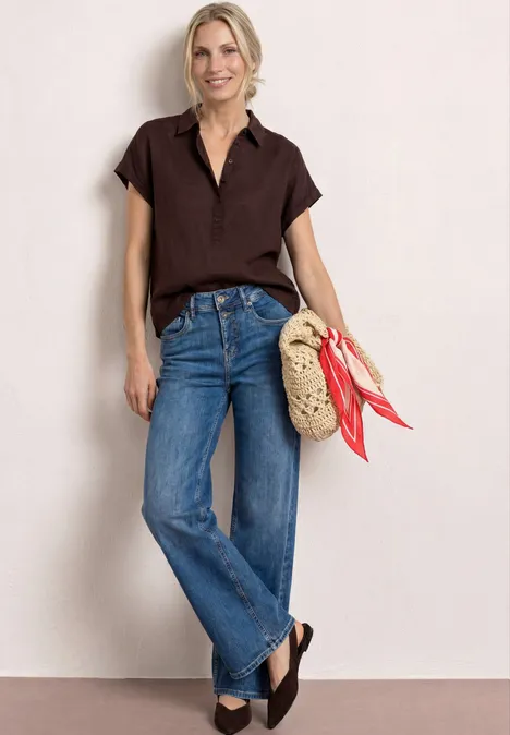 Frau in braunem Kurzarmshirt und blauen Jeans, mit geflochtenem Korbtasche und dunklen Keilabsatzschuhen.