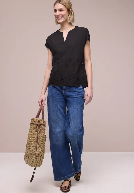 Vrouw met zwarte kanten top, wijdvallende jeans, luipaardballerinas en gevlochten tas tegen een beige achtergrond.