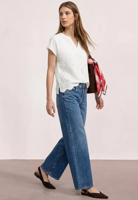 Vrouw in witte korte mouwen blouse en wijde blauwe jeans, zwarte platte schoenen, met een rood sjaaltje.