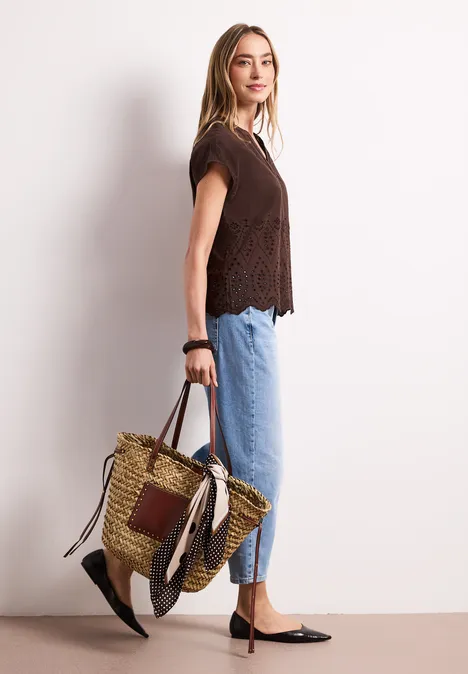 Frau im Profil mit Spitzen-Top, blauen Jeans, schwarzen Ballerinas und geflochtener Strohtasche.