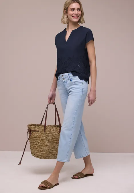 Vrouw in marineblauw shirt, lichtblauwe jeans, bruine sandalen en een grote stroentas voor een neutrale achtergrond.