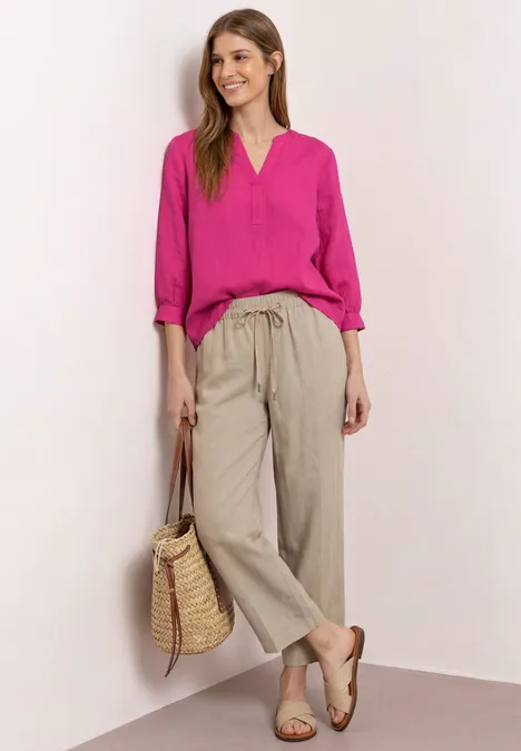 Eine Frau trägt eine pinke Bluse, beige Leinenhose, Sandalen und hält eine geflochtene Strohtasche.