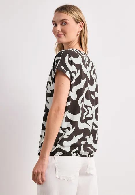 Vrouw in profiel met zwart-wit patroonshirt met korte mouwen en witte broek tegen een neutrale achtergrond.