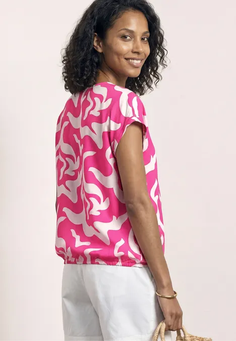 Frau von hinten mit pink-weiß gemustertem Top, weißer Hose und Basttasche.