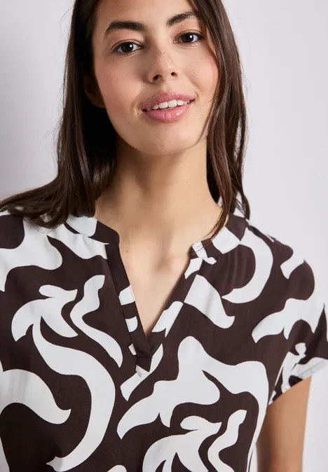 Vrouw in een kort bruin-wit T-shirt met abstract patroon, korte mouwen en V-hals, neutrale achtergrond.