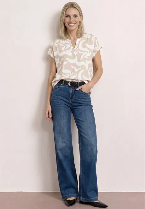 Femme en t-shirt clair à motifs, jean évasé bleu foncé et ceinture, devant un mur neutre.