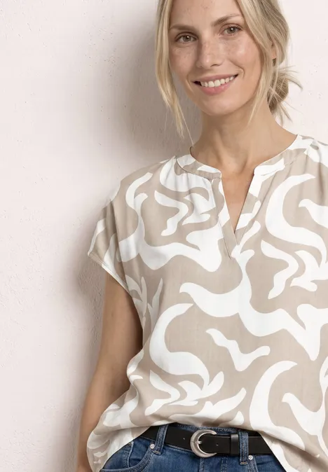 Femme souriante portant une blouse beige à motif blanc et un jean, sur fond neutre.