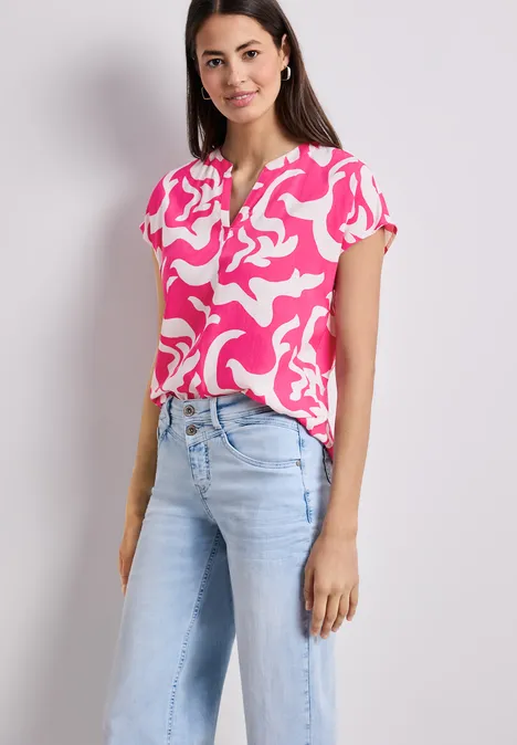 Vrouw draagt roze blouse met witte print en lichte jeans, voor een neutrale achtergrond.