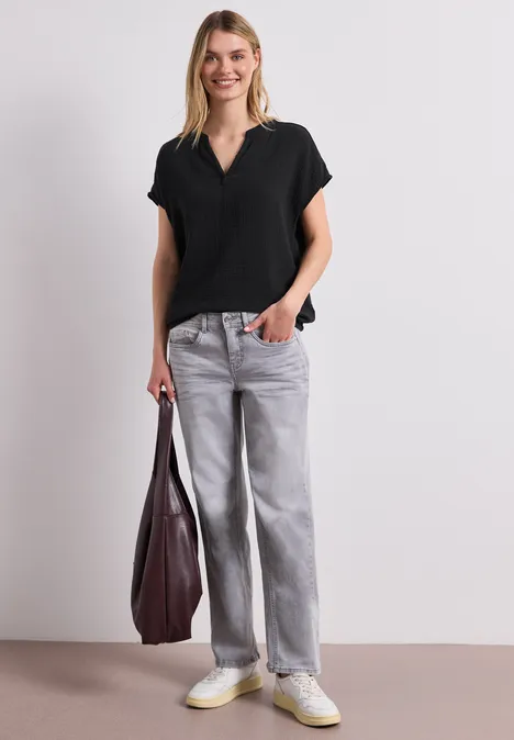 Vrouw in zwarte V-hals top, grijze wijde jeans, witte sneakers, met een grote bordeauxrode tas.