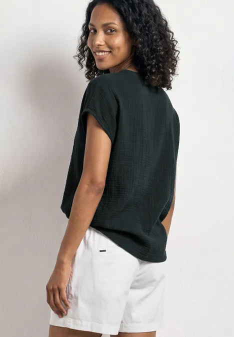 Persoon met krullend haar draagt zwart shirt en witte shorts, staat met de rug naar de camera, opzij gericht.