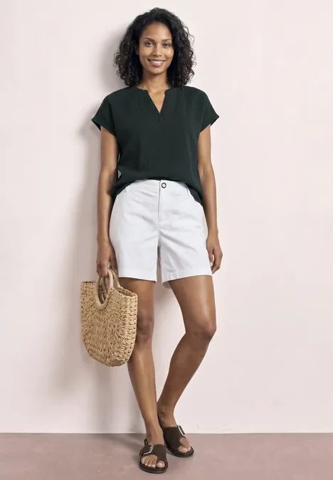 Model draagt zwart T-shirt, witte short, zwarte sandalen en een strooien tas tegen lichte achtergrond.